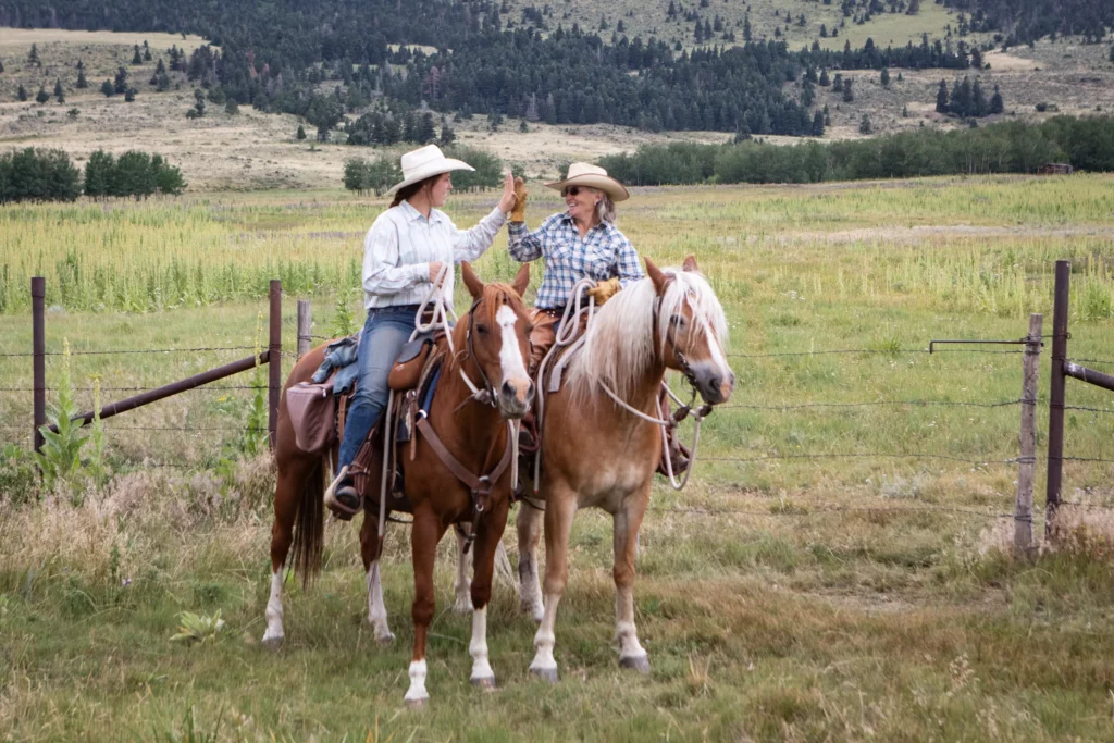 two cowgirls on horseback smiling and giving each other a high five at the Music Meadows Ranch